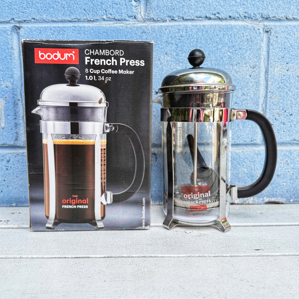 Bodum French press coffee maker next to its packaging against a blue brick wall. 3 cup French press