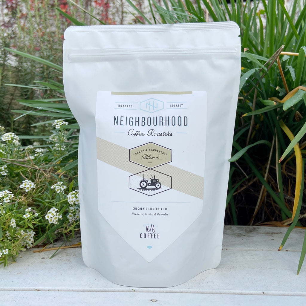 A white bag of Neighbourhood Coffee Roasters organic gardeners coffee with branding and label visible, placed against a garden background.