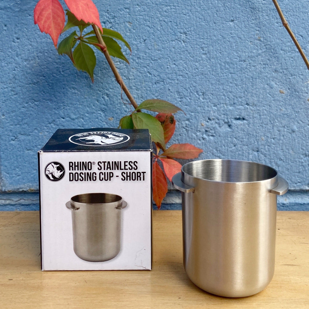 A stainless steel dosing cup next to its packaging box, with a blue background and a red flower in the background.