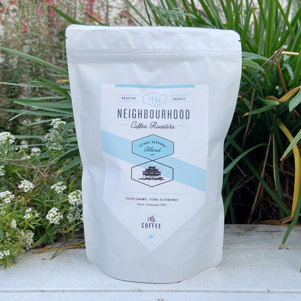 A white bag of Neighbourhood Coffee's Store Keepers Blend coffee, placed against a garden backdrop.
