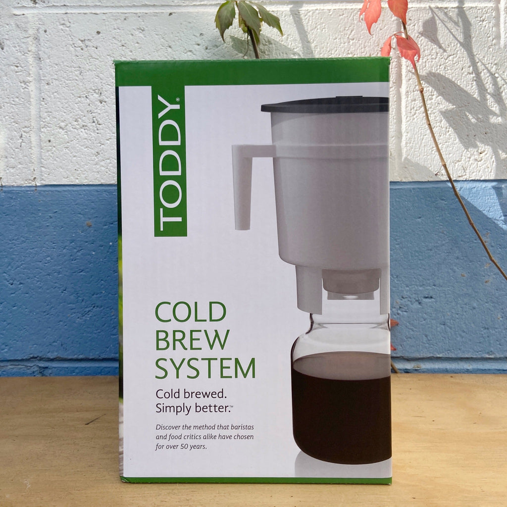 Packaging box of Toddy Cold Brew Coffee Maker with the product visible through a cut-out window. The box is green and white with text and the product image is of a gray and white coffee maker with a glass decanter.