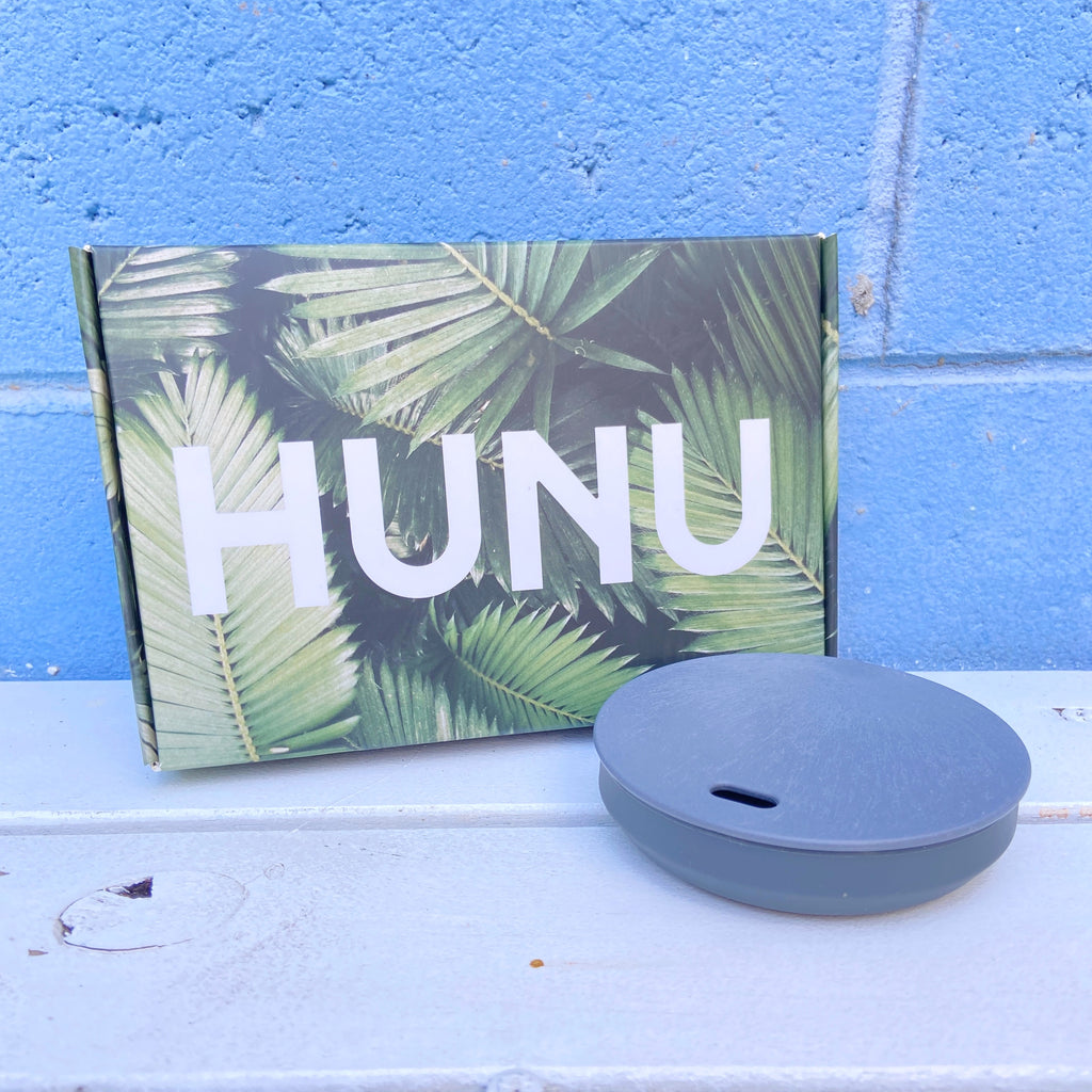 A HUNU collapsible coffee cup in grey, displayed next to its packaging that has a leaf pattern. The cup is made of silicone and appears to be solid gray in color.