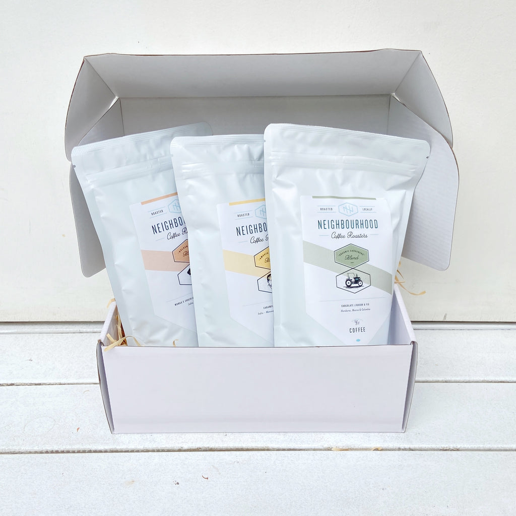 A white glossy gift box open to reveal three sealed bags of coffee beans labeled 'Neighbourhood Coffee Roasters' with different blend names.