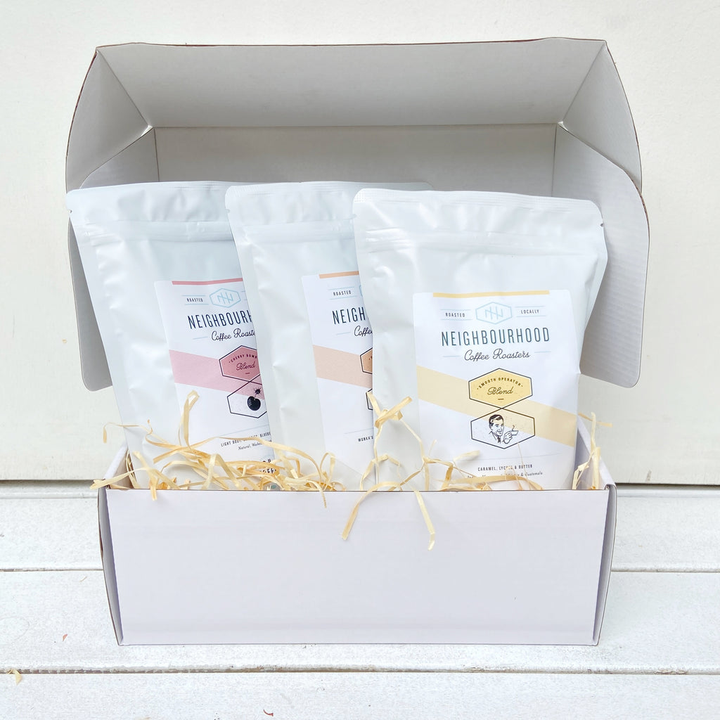 A white glossy gift box open, displaying three white coffee bags labeled Cherry Bomb, Golden Ticket, and Smooth Operator, presented on a bed of straw.